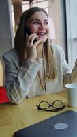 Young lady having pleasant conversation on the phone and smiling. Drinking tea at lunchtime and talking on the smartphone. Cityscape at backdrop. Vertical video