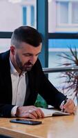 Busy boss writing something in paper notebook. Businessman takes his phone and scrolls information. Big windows at backdrop. Vertical video