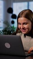 Resilient smiling young woman working on her computer. Happy lady is satisfied with some result and makes a success gesture. Blurred backdrop. Vertical video