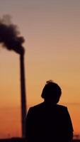 Silhouette of a person with phone at sunset.Man taking pictures of the evening sky on the background of two pipes with dark smoke. Ecological problems. Vertical video