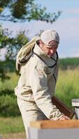 Male apiarist uses a smoker and a brush in his work at apiary. Checking up the bees and honey harvest on sunny summer day. Vertical video