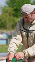 Bearded man in protective outfit taking frames out of hive. Man uses metal tool to extract frames. Blurred nature backdrop. Vertical video