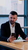 Serious focused businessman uses smartphone sitting at the desk. Man looking for information via phone and taking notes in his paper notebook. Vertical video