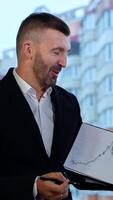 Talkative man shows a graph and comments it to his colleagues. Positive man in official suit explaining charts at office. Big window with house at the backdrop. Vertical video