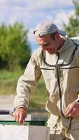 Happy smiling male beekeeper uses metal instrument to extract a frame from hive. Man picks up a frame and shows it to camera. Vertical video