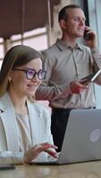 Happy smiling lady works on her laptop at the desk. Man having conversation on the mobile phone at the backdrop. Vertical video