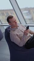 Male worker resting in the bean bag chair. Man searches information on the phone. Panoramic window with city view at backdrop. Vertical video