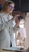 Good-looking lady standing in front of desk and taking notes on paper. Smiling woman makes a phonecall and looks at the screen. Vertical video