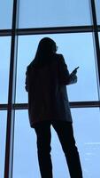 Dark silhouette of a long-haired woman standing with her back. Lady speaking on the phone in front of panoramic window. Low angle view. Vertical video
