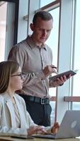 Colleagues have conversation at the table near the window. Woman employee sitting at computer and man standing over her. Vertical video