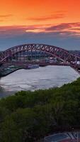 Hell Gate Bridge connecting two green banks. Picturesque structure at the backdrop of orange sky at sun dusk. Vertical video