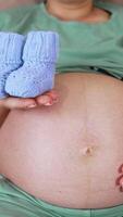 Leaned back pregnant woman on the couch stroking her big belly with one hand. In her other hand she's holding baby booties and shoving them closer to the camera while camera moves back. Vertical video