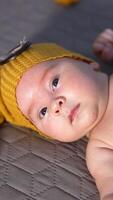 Bare baby in a funny yellow cap lying on the bed. Small boy lying calmly on the bed and look at the camera. Vertical video