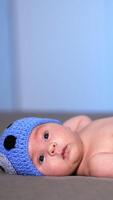 Toddler in a cute knitted cap with ears and diaper lying on the bed. Sweet child looking attentively at the camera. Side view. Vertical video