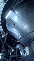 Huge dairy tank beside the thick plastic tubes. Stainless steel tank standing at the raised platform with sensors at its basics. Circle view from below. Vertical video