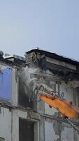 Excavator claw breaks the parts of ruined building. Dangerous deconstruction site with falling debris. Demolishing of the upper floors. Vertical video