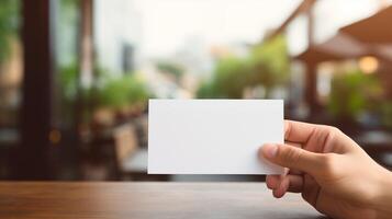 Hand holding blank white card over blurred outdoor background with greenery. Cafe table. Branding, identity template advertising image. Promotional design mock up photorealistic photo