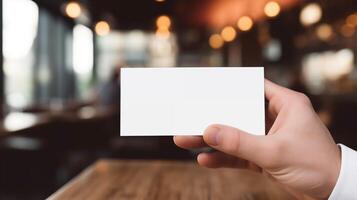 Hand holds blank white business card mockup in warm indoor cafe. Restaurant setting template advertising image. Blurred lights, cozy ambiance. Identity mock up product photorealistic photo