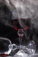 Two glasses on a black background. Smoke around a glass of wine. A glass of red wine stands on the surface, an empty glass lies next to it. photo