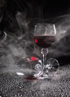 Two glasses on a black background. Smoke around a glass of wine. A glass of red wine stands on the surface, an empty glass lies next to it. photo