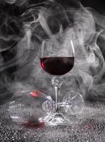 Two glasses on a black background. Smoke around a glass of wine. A glass of red wine stands on the surface, an empty glass lies next to it. photo