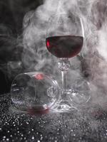 Two glasses on a black background. Smoke around a glass of wine. A glass of red wine stands on the surface, an empty glass lies next to it. photo