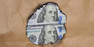 A portion of a US bill featuring Benjamin Franklin, the bill visible through a burnt hole in the brown paper photo