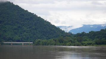 tranquil riverside landscape featuring a green, forested mountain with a bridge crossing the river. The misty background and cloudy skies add an atmospheric, peaceful tone video