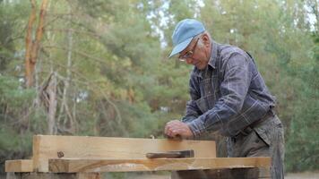 An experienced craftsman carefully processes a wooden board using an old plane. Surrounded by tall trees and natural light, he focuses on his work and enjoys the manual labor. video