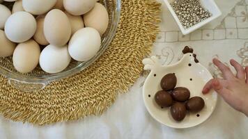 A child reaches for a chocolate egg from a chicken-shaped dish on Easter. is shot from a top-down perspective. The table is covered with a white tablecloth, and bowl of white and brown eggs. video