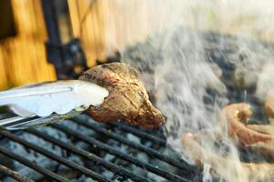 Close-up of tongs lifting a sizzling steak from a hot barbecue grill, with smoke rising, capturing the essence of outdoor grilling photo