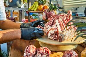 A chef wearing black gloves arranges a rack of raw lamb on a wooden board surrounded by various cuts of meat, preparing for cooking. photo