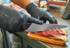 A close-up of a chef's hands in black gloves using a knife to slice raw meat on a small wooden cutting board, preparing for cooking photo