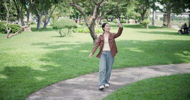 Handheld shot, Cheerful young woman holding a smartphone and chatting happily with her friend via call while walking in a park. A holiday time surrounded by nature video