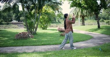 Handheld back view shot, Happy woman enjoys takes a photo on mobile phone while walking in a park. A holiday time surrounded by nature video
