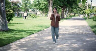 Handheld shot, Happy woman enjoys talking on her mobile phone while walking in a park. A holiday time surrounded by nature video
