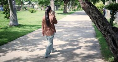 Handheld back view shot, Happy woman enjoys talking on her mobile phone while walking in a park. A holiday time surrounded by nature video
