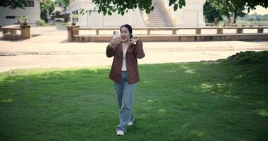 Handheld shot, Cheerful young woman holding a smartphone and chatting happily with her friend via call while walking in a park. A holiday time surrounded by nature video