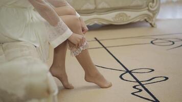 Legs of bride in night gown and veil. Sitting on a sofa and puts garter on a leg video