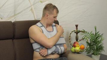 Painful, bored man with a broken arm wearing arm brace sitting on a sofa video