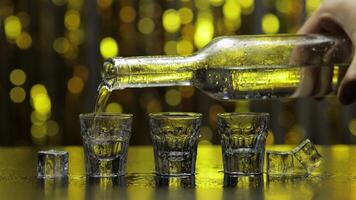 Barman pour frozen vodka from bottle into shot glass. Ice cubes against shiny gold party background video