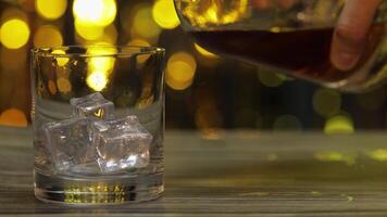 Pouring of golden whiskey, cognac or brandy from bottle into glass with ice cubes. Shiny background video