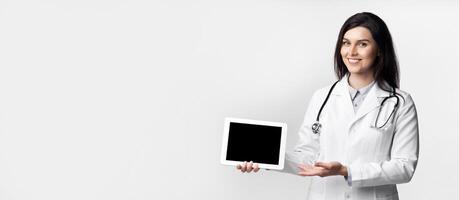A female doctor wearing a white lab coat and stethoscope smiles while holding a tablet with a blank screen. photo