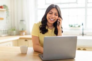 A middle eastern woman is multitasking, talking on her cell phone while working on her laptop. She appears focused and engaged in a conversation while typing on the computer. photo
