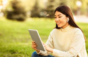 Asian woman is seated on the grass, engaged with a tablet device. She appears focused on the screen, possibly reading or watching something. The grass is green and well-kept photo