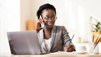 Smiling African American Secretary Talking On Phone And Taking Notes While Working On Laptop In Modern Office, Free Space photo