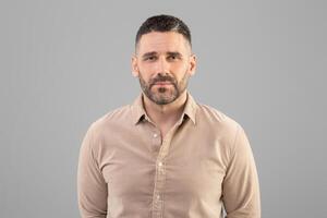 A man with a short haircut and beard is standing against a plain gray background, wearing a beige shirt and looking directly at the camera. The setting appears to be indoors with even lighting. photo