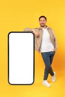 A smiling man in casual attire leans confidently against an oversized, blank white screen of a smartphone model, mockup copy space photo