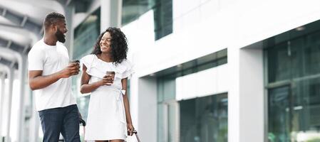 Black couple walks side-by-side on the sidewalk outside a modern building. They both have coffee cups in their hands and are smiling as they talk to each other, panorama with copy space photo