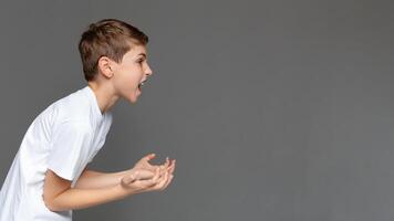 Awkward age. Angry emotional boy screaming and gesturing with hands in air, side view, gray studio background with free space photo